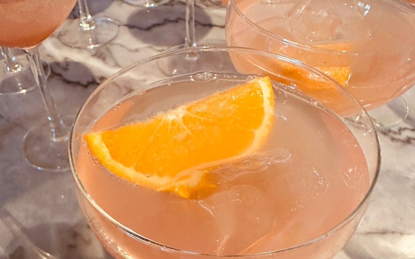 Roze-oranje spritz met een schijfje sinaasappel in coupeglazen op een marmeren tafel